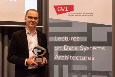 Marcin Zukowski with the Fellowship Award standing next to a banner announcing lectures on Data Systems Architectures